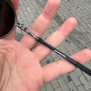 May include: Black sunglasses with dark lenses and a thin, dark metal frame. The temple arm features the white text "MAD". The sunglasses are held in a hand against a gray brick background.