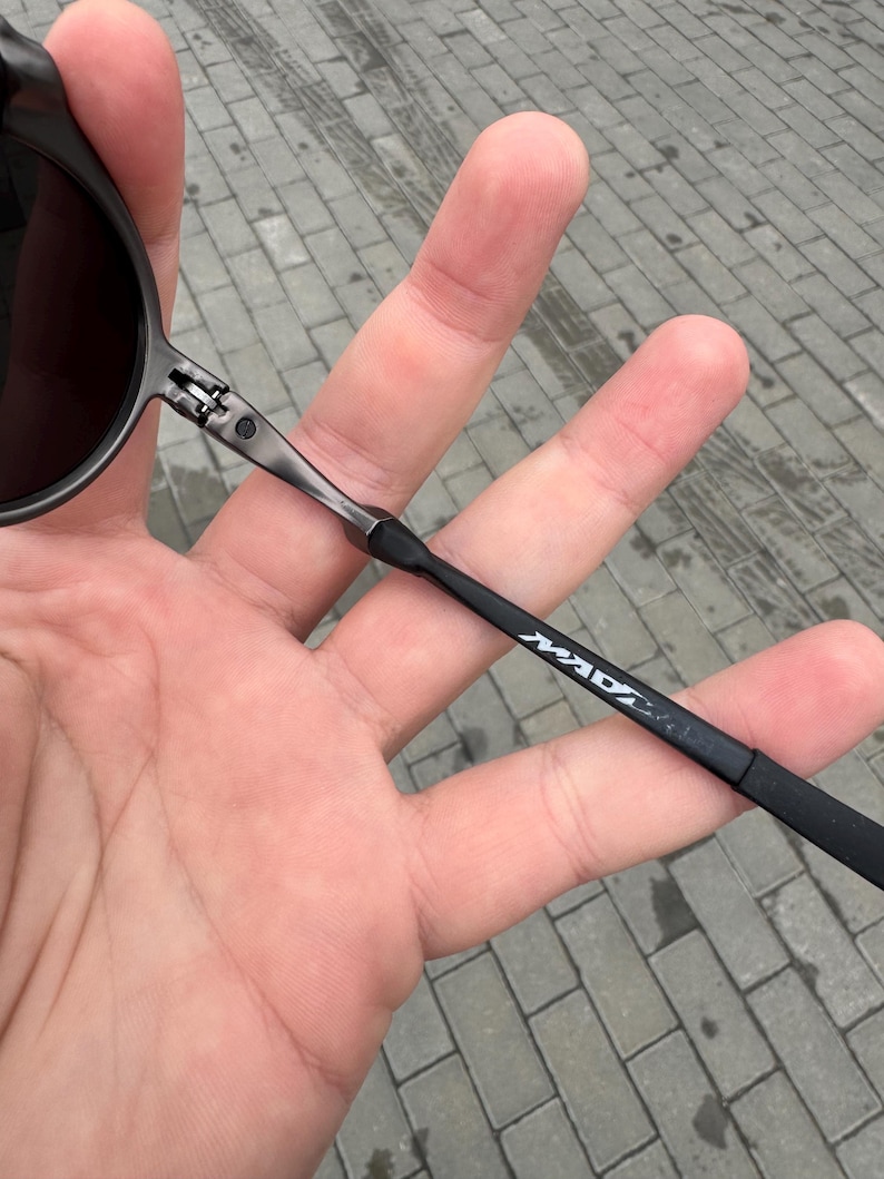 May include: Black sunglasses with dark lenses and a thin, dark metal frame. The temple arm features the white text "MAD". The sunglasses are held in a hand against a gray brick background.