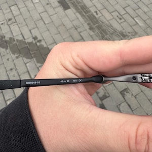 May include: Black eyeglasses with the text "OO6019-01" and measurements "42-29 151 CE" printed on the arm. The glasses are held in a hand against a gray brick background.