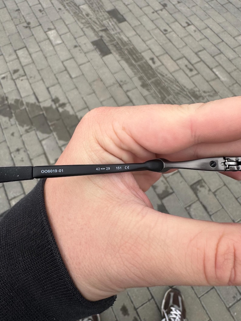 May include: Black eyeglasses with the text "OO6019-01" and measurements "42-29 151 CE" printed on the arm. The glasses are held in a hand against a gray brick background.