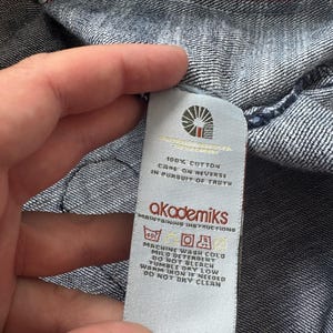 Może przedstawiać: Zbliżenie na dżinsową odzież z białą metką. Etykieta zawiera nazwę marki "akademiks" i instrukcje pielęgnacji. Tkanina to niebieski dżins z czerwonymi szwami.