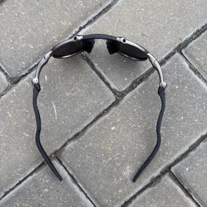 May include: A pair of sunglasses with a black frame and dark lenses. The arms are silver and curve down to black ear pieces. The sunglasses are lying on a gray brick surface.