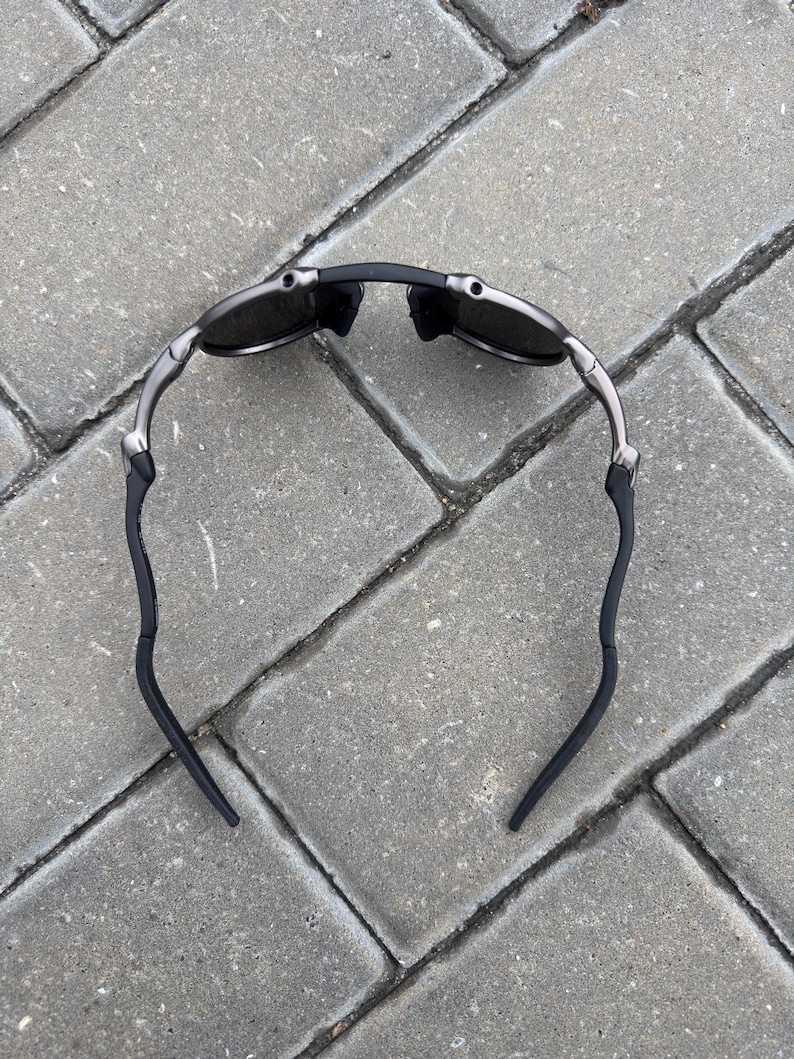 May include: A pair of sunglasses with a black frame and dark lenses. The arms are silver and curve down to black ear pieces. The sunglasses are lying on a gray brick surface.
