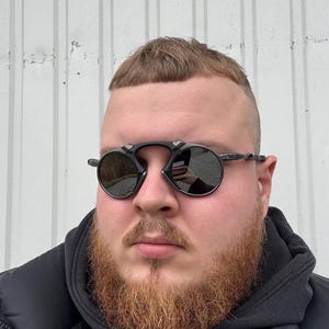May include: Black round sunglasses with a dark tint. The sunglasses have a thin frame and are worn by a person with a red beard and short hair. The background is a white wall.