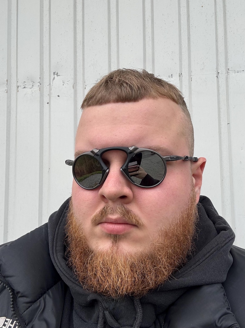 May include: Black round sunglasses with a dark tint. The sunglasses have a thin frame and are worn by a person with a red beard and short hair. The background is a white wall.