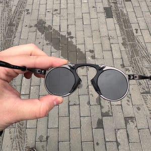 May include: Round sunglasses with a black frame and dark lenses. The sunglasses have a unique bridge design and thin temples. The lenses are circular and appear to be polarized. The sunglasses are held up against a gray brick background.