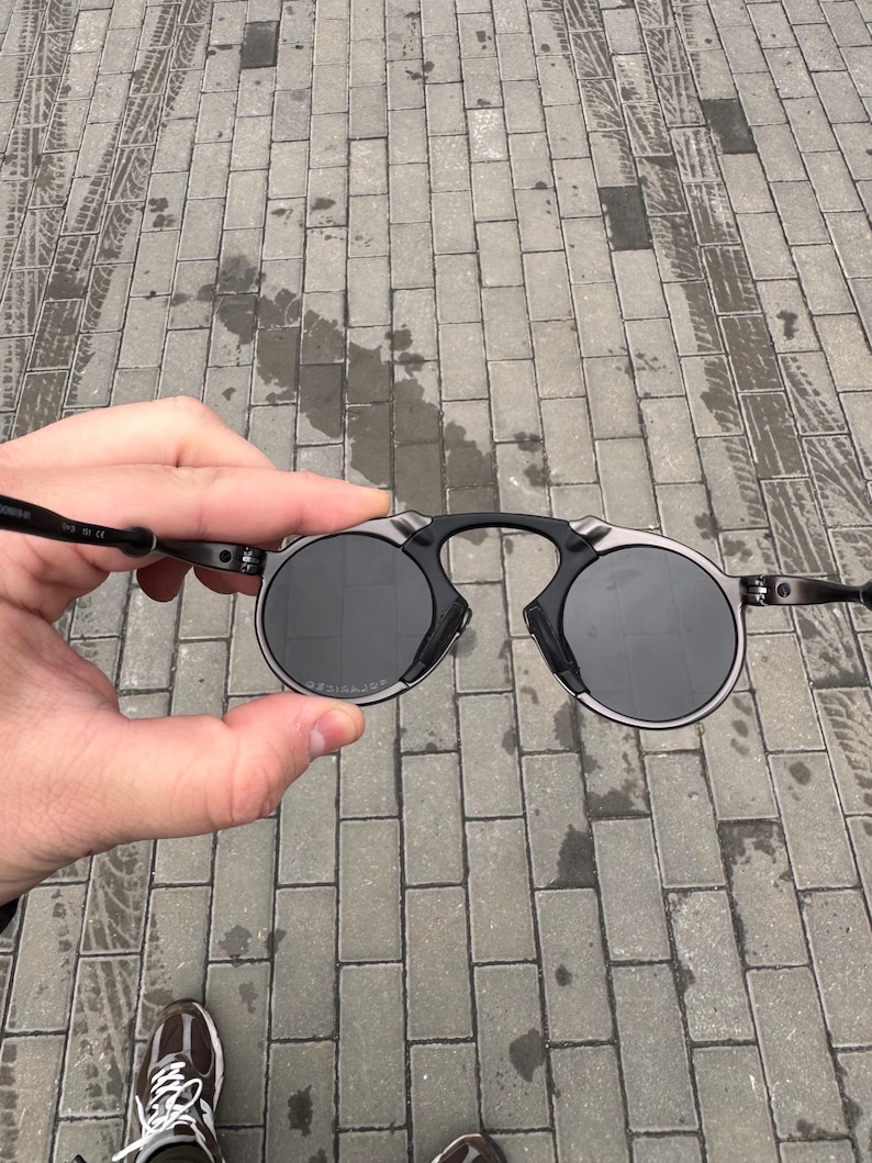 May include: Round sunglasses with a black frame and dark lenses. The sunglasses have a unique bridge design and thin temples. The lenses are circular and appear to be polarized. The sunglasses are held up against a gray brick background.