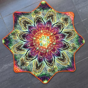 Op de afbeelding: Een stervormige mat met een levendig, veelkleurig mandalapatroon in tinten paars, geel, groen en oranje. Het midden toont een lichtblauw geometrisch knoopsymbool en de gehele rand is afgewerkt met een feloranje bies.