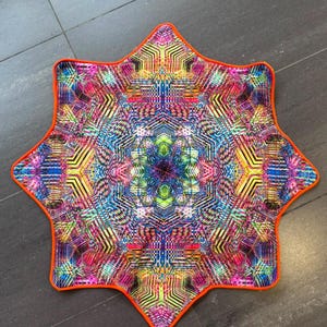 Könnte beinhalten: Eine sternförmige Matte mit einem lebendigen, kaleidoskopischen Muster aus komplexen geometrischen Linien in Neonrosa, Elektroblau, leuchtendem Gelb und Dunkelviolett, eingefasst mit einer soliden, orangefarbenen Stoffkante.