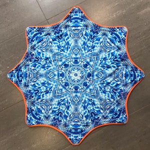 Op de afbeelding: Een blauw en wit stervormig tapijt met een ingewikkeld mandala-ontwerp. Het tapijt heeft een oranje rand.