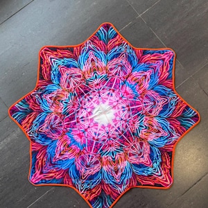 Op de afbeelding: Een stervormige, doorgestikte mat met een levendig, symmetrisch mandalapatroon in neonkleuren zoals roze, blauw en paars. Het stoffen oppervlak toont complexe, lichtgevende bloemmotieven die vanuit het midden uitstralen, afgewerkt met een feloranje rand.