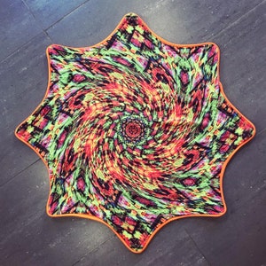 Może przedstawiać: Kolorowy dywan w kształcie gwiazdy z wirującym, psychodelicznym wzorem w odcieniach czerwieni, pomarańczy, żółci, zieleni i błękitu. Dywan ma pomarańczową obwódkę.