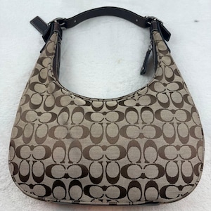 Könnte beinhalten: Eine Schultertasche im Hobo-Stil mit einem beigen und dunkelbraunen Monogramm-Muster. Die Tasche verfügt über einen einzelnen dunkelbraunen Lederriemen mit silberfarbenen Metallschnallen, dunkelbraune Lederbesätze und einen Reißverschluss.