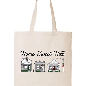 May include: A white canvas tote bag with the text "Home Sweet Hill" printed in black cursive font. Three small houses are illustrated below the text, each with a different design and color.