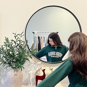 May include: A dark green quarter-zip sweatshirt with the text "THE HILL" in white lettering. The sweatshirt is reflected in a large round black mirror. Decorative items, including a vase with greenery and deer figurines, are on a white surface.
