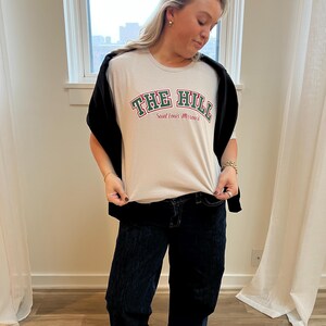 May include: A light beige t-shirt with the text "THE HILL" in green and red lettering, with "Saint Louis Missouri" below. The shirt is paired with dark blue jeans and a black sweater.
