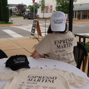 May include: A tan t-shirt and a white cap with the text "ESPRESSO MARTINI D'Italia COCKTAIL CLUB" and "IL DOLCE Far Niente" in black. A black cap with the same text is also visible. The items are on a white table.