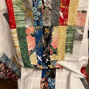 May include: A patchwork quilt with a variety of patterned fabrics in red, gold, green, blue, and grey. The fabrics include floral, crane, and geometric designs. The quilt is bordered with white fabric.