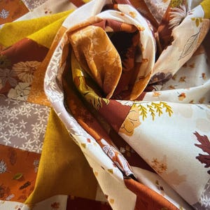May include: A close-up of a fabric quilt with a patchwork design featuring various shades of brown, yellow, and white. The fabric is patterned with leaves and flowers.