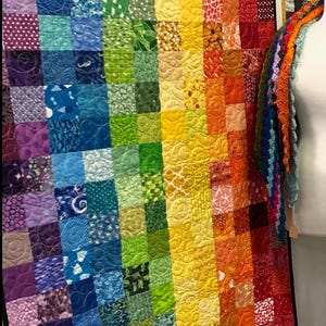 Könnte beinhalten: Eine lebendige, handgefertigte Steppdecke, die an einer weißen Wand ausgestellt ist. Die Steppdecke zeigt einen Regenbogen an Farben, wobei jede Reihe einen anderen Farbton zeigt. Die Quadrate bestehen aus verschiedenen gemusterten Stoffen und schaffen ein visuell ansprechendes Textilkunstwerk.