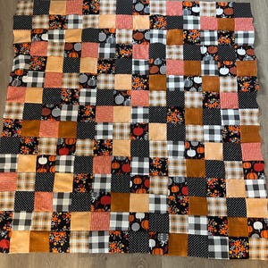 May include: A patchwork quilt featuring a variety of autumn-themed fabrics. The quilt is composed of squares in shades of orange, brown, black, and white. Patterns include pumpkins, polka dots, plaid, and floral designs. The quilt is a rectangular shape.