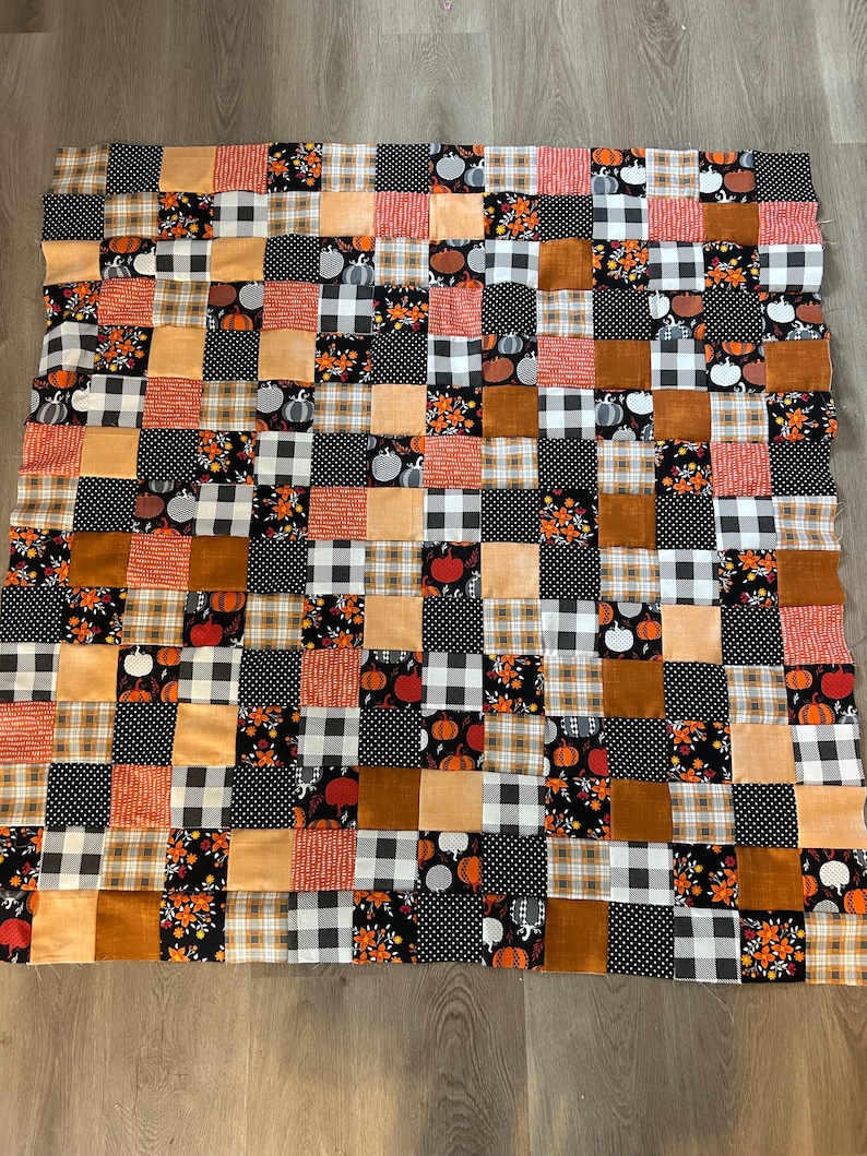 May include: A patchwork quilt featuring a variety of autumn-themed fabrics. The quilt is composed of squares in shades of orange, brown, black, and white. Patterns include pumpkins, polka dots, plaid, and floral designs. The quilt is a rectangular shape.