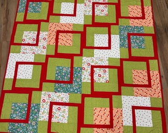 Op de afbeelding: Een rechthoekige quilt met een geometrisch patroon van in elkaar grijpende vierkanten en L-vormige segmenten. Het ontwerp combineert effen limoengroene stof met bloemenprints in wit, perzik en blauwgroen, omlijst door helderrode randen.