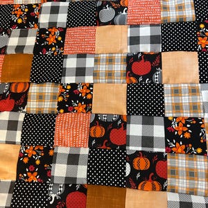 May include: A patchwork quilt featuring a variety of autumn-themed fabrics. The quilt incorporates squares of orange, black, and tan fabrics with patterns including pumpkins, polka dots, plaid, and floral designs. The quilt is a cozy, decorative item.