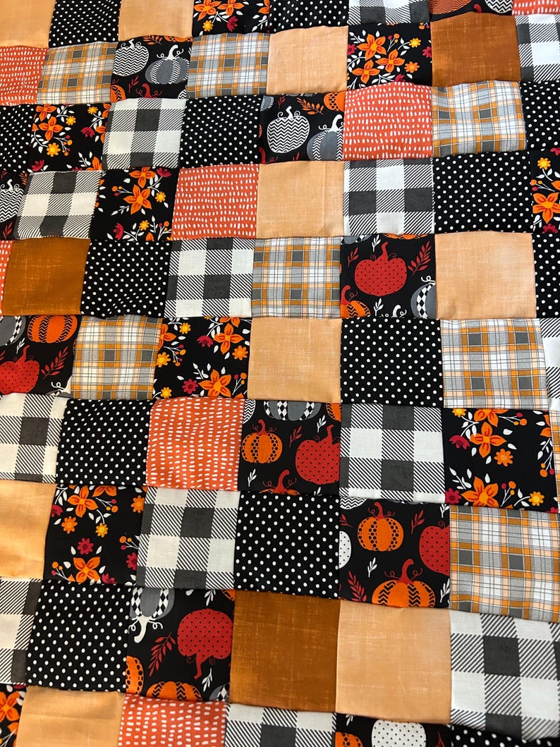 May include: A patchwork quilt featuring a variety of autumn-themed fabrics. The quilt incorporates squares of orange, black, and tan fabrics with patterns including pumpkins, polka dots, plaid, and floral designs. The quilt is a cozy, decorative item.