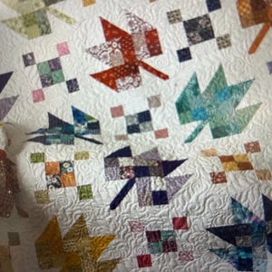 May include: A white quilt featuring a leaf design. The leaves are crafted from diverse patterned fabrics in red, green, yellow, and blue hues. Small square fabric blocks are interspersed around the leaves. The quilt has a quilted, swirling pattern.
