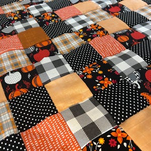 May include: A patchwork quilt with a fall theme. The quilt features squares of various fabrics in orange, black, white, and tan. Patterns include polka dots, plaid, floral designs, and pumpkin motifs. The quilt is a cozy and decorative home textile.