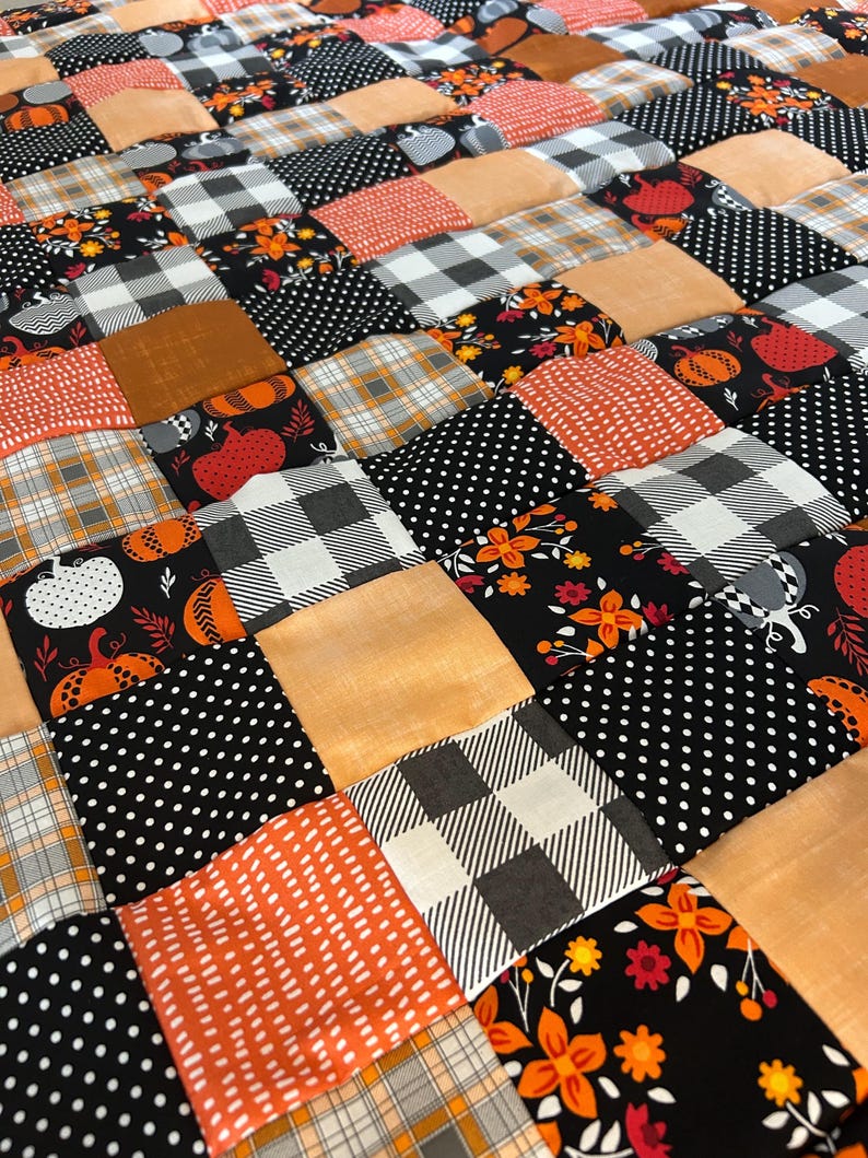 May include: A patchwork quilt with a fall theme. The quilt features squares of various fabrics in orange, black, white, and tan. Patterns include polka dots, plaid, floral designs, and pumpkin motifs. The quilt is a cozy and decorative home textile.