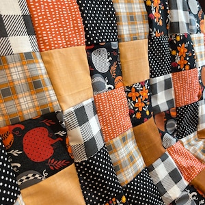 May include: A patchwork quilt with a fall theme. The quilt features squares of fabric in various patterns, including pumpkins, plaid, polka dots, and floral designs. The color palette includes orange, black, white, and gray.