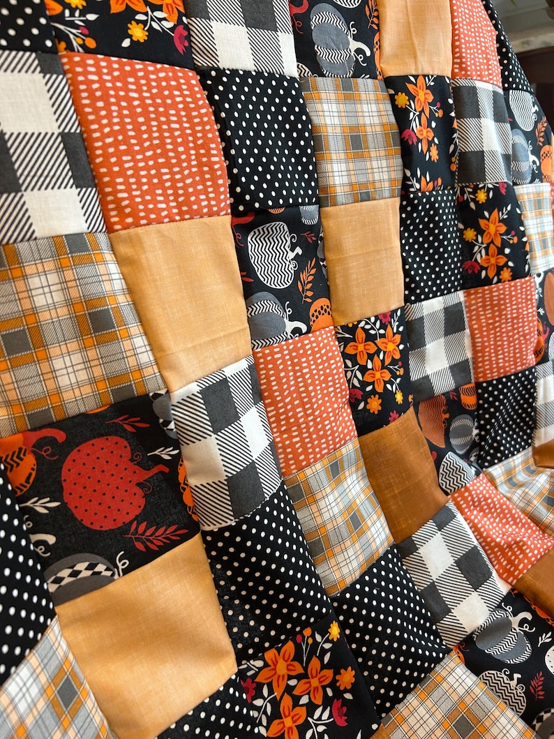 May include: A patchwork quilt with a fall theme. The quilt features squares of fabric in various patterns, including pumpkins, plaid, polka dots, and floral designs. The color palette includes orange, black, white, and gray.