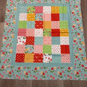 Puede incluir: Un edredón de retazos con un borde floral y un cuadrado central de cuadrados coloridos. Los cuadrados presentan varios patrones, incluyendo cuadros, fresas y estampados florales. El borde es azul claro con flores rojas y rosas.