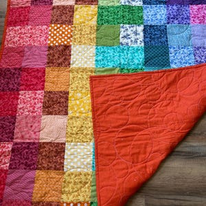 May include: A handmade patchwork quilt with a vibrant rainbow color scheme. The quilt features squares of various patterned fabrics, including polka dots and floral designs. The underside is a solid orange with a circular quilted pattern.