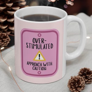 Könnte beinhalten: Ein weißer Keramikbecher mit einem rosa rechteckigen Schildmotiv mit abgerundeten Ecken. Der Text lautet OVER-STIMULATED, gefolgt von einem gelben Dreieck mit Ausrufezeichen und dem schwarzen Text APPROACH WITH CAUTION.