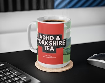 Può includere: Una tazza di ceramica bianca con la scritta "ADHD & YORKSHIRE TEA" in un design rosso e bianco. La tazza è riempita con un liquido scuro, probabilmente caffè o tè, e poggia su un sottobicchiere di sughero. Il vapore sale dalla tazza.