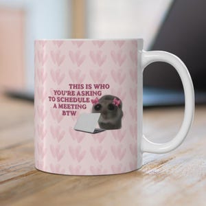 Könnte beinhalten: Ein weißer Keramikbecher mit einem sich wiederholenden hellrosa Herzmuster. In der Mitte befindet sich eine Grafik eines kleinen grauen Wesens mit rosa Schleifen auf dem Kopf, das einen Laptop benutzt, mit dem Text: THIS IS WHO YOU'RE ASKING TO SCHEDULE A MEETING BTW.