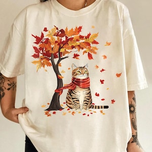 May include: An off-white short-sleeve t-shirt featuring a graphic of a brown tabby cat wearing a red and white plaid scarf. The cat sits under a tree with orange, yellow, and red autumn leaves, with several leaves falling around the cat.