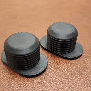 May include: Two black, round, plastic bottle stoppers with a ridged design. The stoppers are sitting on a brown surface.