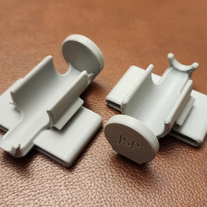 May include: Two gray plastic 3D printed clips with a round base. The base of one clip has the letters "JSP" printed on it.