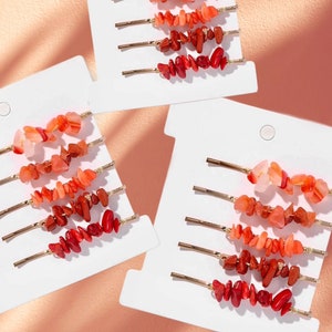 Puede incluir: Conjunto de 12 pinzas para el pelo doradas con detalles de piedras rojas, naranjas y rosas. Las pinzas están dispuestas en tarjetas blancas.