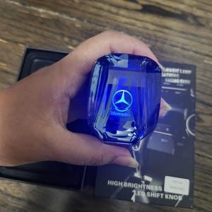 May include: A blue, translucent, crystal-style gear shift knob featuring an illuminated Mercedes logo and text. The product is displayed with its black packaging box, which includes the printed text: HIGH BRIGHTNESS LED SHIFT KNOB.