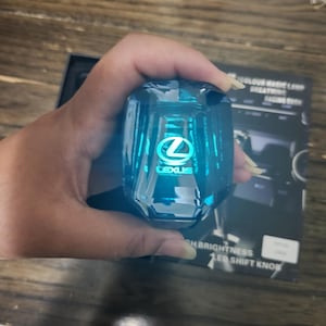 May include: A blue, faceted LED shift knob with the Lexus logo illuminated in the center. The knob is held in a hand, with the packaging visible in the background. The packaging includes the text "HIGH BRIGHTNESS LED SHIFT KNOB".