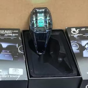 May include: A clear, faceted gear shift knob with internal LED lighting, displayed in a black foam-lined box. The packaging features the text "MAGIC LAMP BREATHING RACING DASH" and a Toyota logo, with additional branding for M-spirit LED 70.