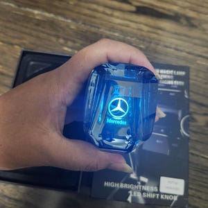 May include: A blue, illuminated Mercedes-Benz LED gear knob. The knob is held in a hand and has the Mercedes logo and text. The packaging includes the words "High Brightness LED Shift Knob".