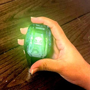 Puede incluir: Un llavero de coche Toyota verde brillante. El llavero se sostiene en una mano y tiene el logotipo de Toyota visible. El llavero tiene forma ovalada y tiene una base metálica.