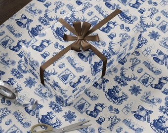 Puede incluir: Un rollo de papel de regalo navideño y un regalo envuelto con una cinta marrón sobre una mesa de madera. El papel presenta un patrón toile de Jouy azul y blanco con ilustraciones festivas, incluyendo campanas, calcetines y renos.