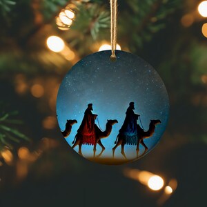 May include: Round Christmas ornament featuring a starry night sky with three silhouetted figures riding camels. The figures are wearing robes of red and blue. A jute string is attached for hanging.